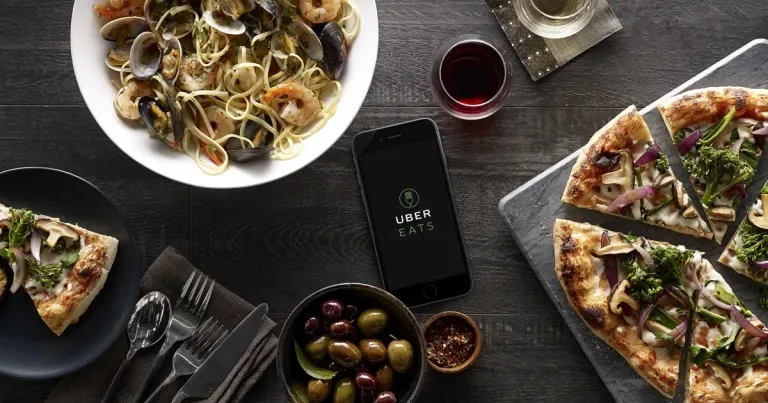 Uber Eats app open on an iPhone on a dark wooden table, surrounded by seafood pasta, pizza, olives and red wine